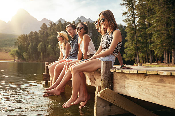 Friends enjoying a day at the lake Portrait of group of young people sitting on the edge of a pier, outdoors in nature. Friends enjoying a day at the lake. jetty stock pictures, royalty-free photos & images
