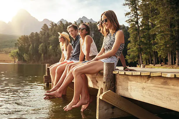 Portrait of group of young people sitting on the edge of a pier, outdoors in nature. Friends enjoying a day at the lake.