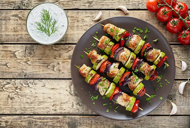 Turkey or chicken meat shish kebab skewers with tzatziki sauce, chopped parsley, garlic and tomatoes on rustic wooden table background. Traditional barbecue grill food