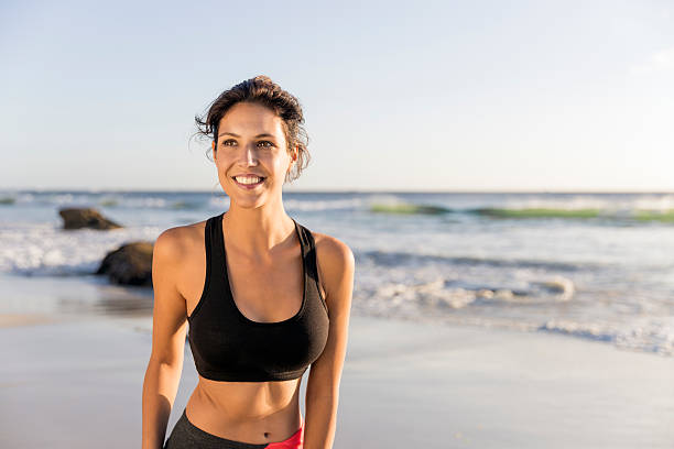 Sporty young woman smiling at beach A photo of fit young woman looking away. Happy female athlete is at beach. She is wearing sportswear while standing against sea. sports bra stock pictures, royalty-free photos & images