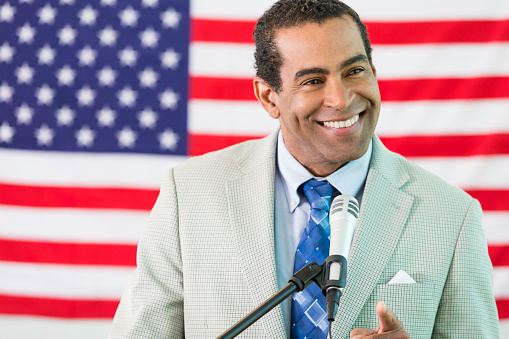 African American man standing in front of the American flag. He is wearing a suit and tie. He is speaking at a microphone.