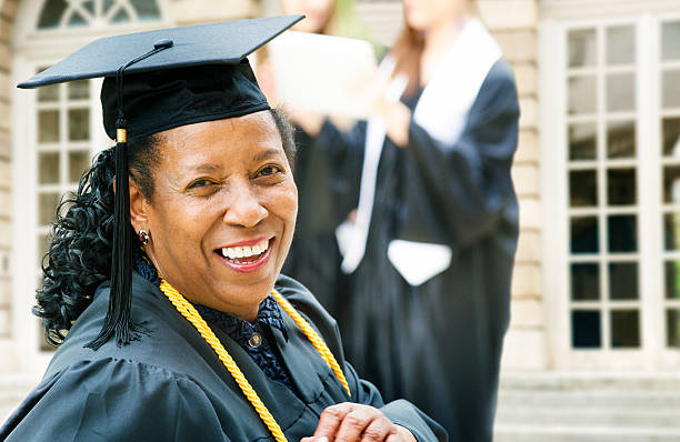Mature African American woman on graduation day Mature African American woman on graduation day, turning to the camera with a smile. Younger graduates are coming down the stairs behind her. nontraditional student photos stock pictures, royalty-free photos & images