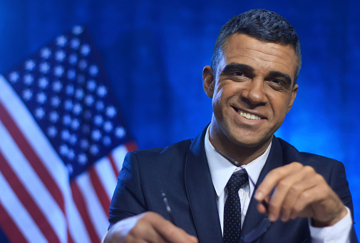 Low angle view at the serious young American politician looking away during his speech at the debates, standing behind the pedestal on a blue background with two American flags