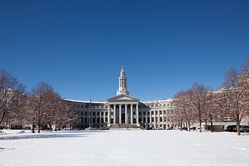 On a clear winter day, snow covers Civic Center Park and the Denver City and County Building under a beautiful blue sky.