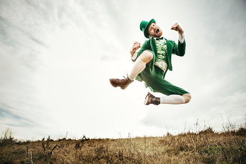 A stereotypical Irish character all ready for Saint Patricks day jumps and dances in an open field of Irish country side.  Copy space in the sky and grass.