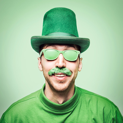 A man smiles, wearing all green clothes, glasses, and even a green mustache in celebration of St. Patty's day.  Square crop with green background.