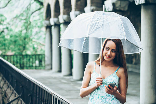 jeune femme tenant un parapluie et à l'aide de téléphone sous la pluie - rain women umbrella parasol photos et images de collection