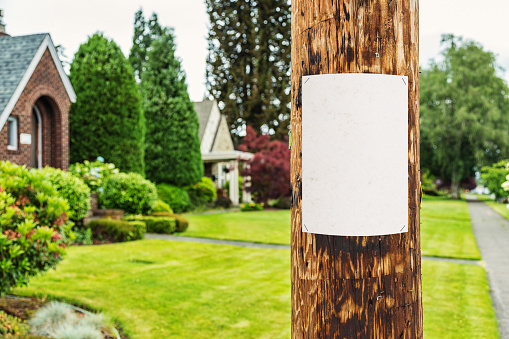 Photo of a blank sheet of white paper fastened to a wooden electrical pole, neighborhood houses visible in the background.