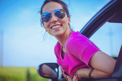 Cheerful, beautiful, young woman rests looking out the car window, enjoying the view and a fresh air. Freedom, travel, vacation and summer road trip concept. Blue sky in the background as a copy space.