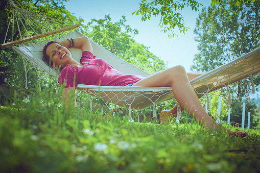 Young carefree woman relaxes in a hammock in the garden, on a bright sunny day. She smiles while napping and daydreaming. The image is taken from the low angle, and there are green leafy trees and a clear blue sky in the background.