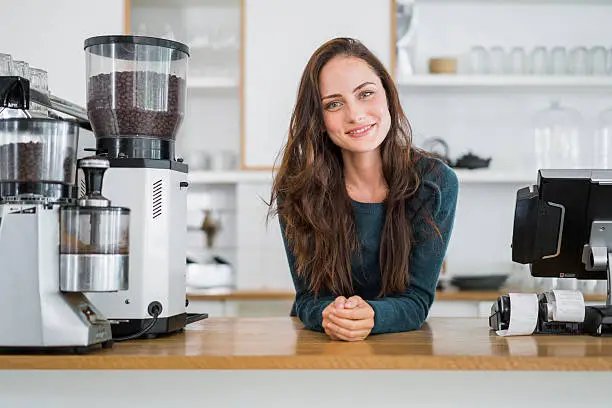 Portrait of confident barista leaning on counter. Attractive female is with long brown hair. She is standing by coffee grinder in cafe.