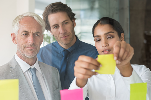 Multi ethnic young businesswoman sticking adhesive notes on glass wall in office. Close up of leadership and his business team looking at sticky notes on window. Business teamwork during a meeting in a board room.