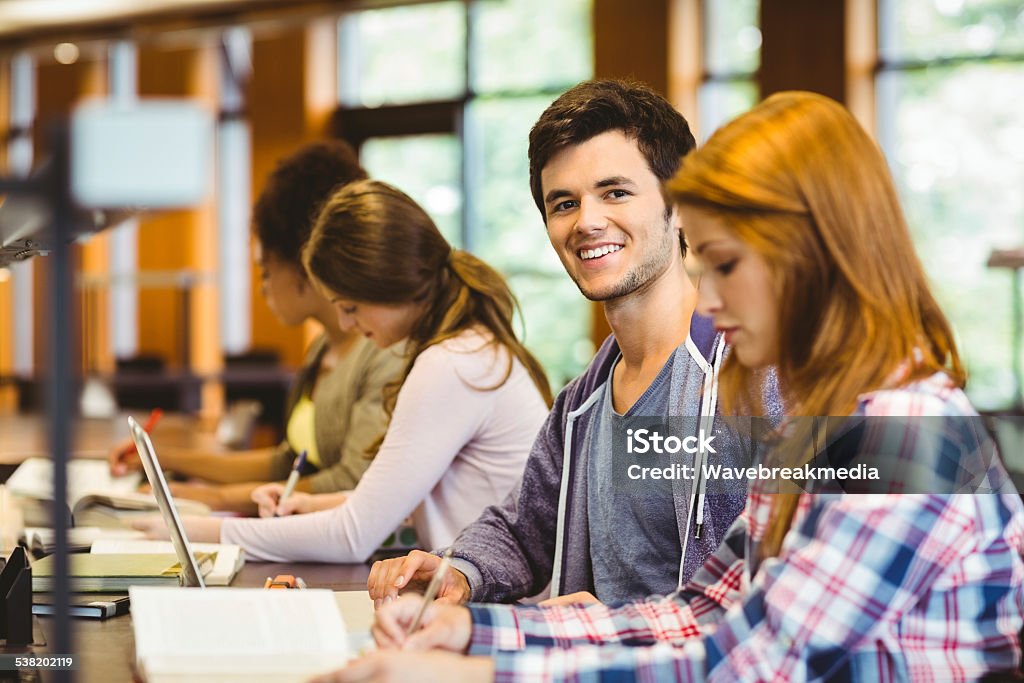 Student looking at camera while studying with classmates Student looking at camera while studying with classmates in library Library Stock Photo