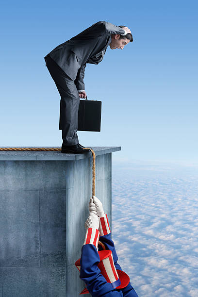 U.S. Government Crisis Uncle Sam hanging from a rope off the side of a building as a businessman helplessly looks on. The businessman is holding a briefcase with his hand on top of his head as he looks down at Uncle Sam. uncle sam broke stock pictures, royalty-free photos & images