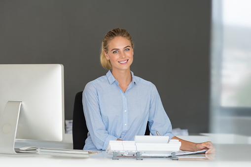 Young beautiful business woman with computer in office. Portrait of beautiful receptionist working on computer in a workplace. Portrait of smiling businesswoman sitting at help desk and looking at camera.