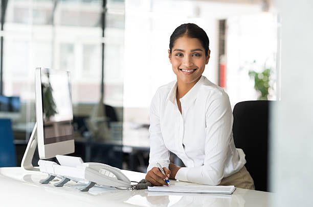 Cheerful woman in office Portrait of beautiful young businesswoman sitting at her desk in front of computer and taking notes. Multi ethnic receptionist looking at camera. Smiling multiethnic business woman working in office. secretary stock pictures, royalty-free photos & images