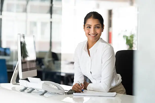 Portrait of beautiful young businesswoman sitting at her desk in front of computer and taking notes. Multi ethnic receptionist looking at camera. Smiling multiethnic business woman working in office.