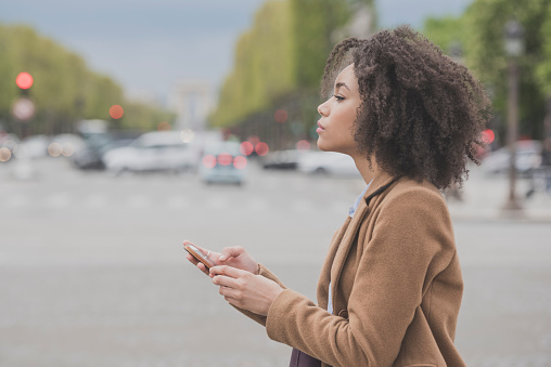 Candid portrait of attractive mixed race female on the street with cell phone, looking away. Side view of woman holding phone. Female tourist using phone in Paris, France