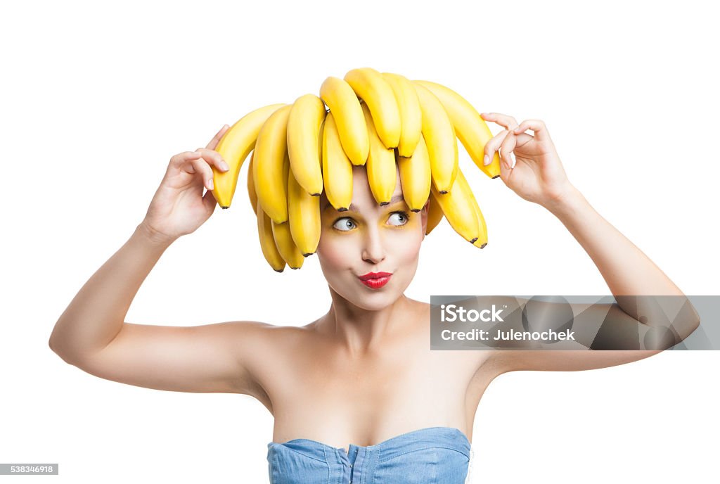 Excited model with bananas on head holding one in hand Happy model with make-up and bunch on bananas on head holding on in hand and looking away. Banana Stock Photo