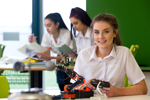 Adolescent girl in a design and technology lesson. She is smiling at the camera with a robotic arm that she is building infront of her.