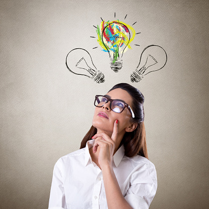 Front shot of a young businesswoman standing and looking up at glowing light bulb over her head as symbol Thinking Outside The Box.