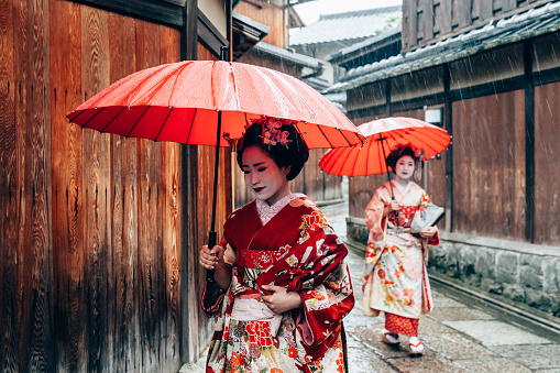 Two maiko geisha walking on a street in Kyoto, Japan during a storm with red umbrella