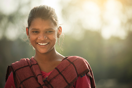 Outdoor image of a happy rural Indian girl giving toothy smile and looking at camera. She is in traditional dress which is Salwar Kameez and Dupatta.  One person, horizontal composition with selective focus.