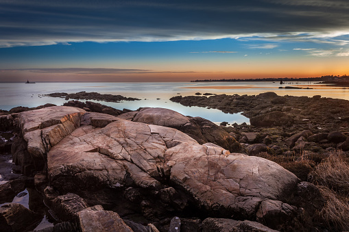 Large rocks on the shore under a golden and blue sunset with a lighthouse in the background.