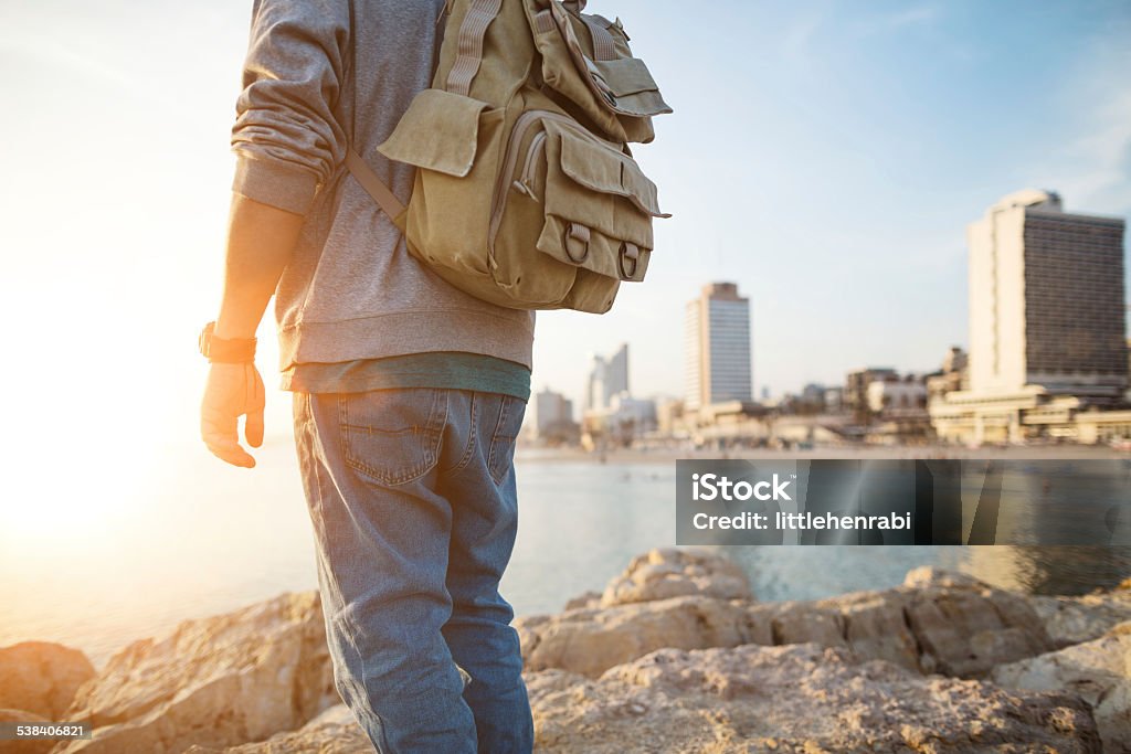 traveler standing on the rocks near the sea traveler with backpack standing on the rocks on the beach in the city and looking far away Backpack Stock Photo