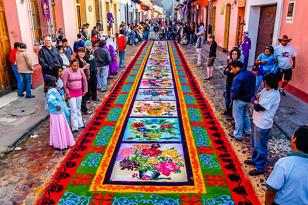 Antigua, Guatemala - April 10, 2009: Spectators admire Holy Week carpet on Good Friday morning in Spanish colonial town & UNESCO World Heritage Site with most famous Holy Week celebrations in Latin America. Groups work all night to make carpet (using wooden stencils and dyed sawdust) in path of Good Friday procession. Purple clad 'cucuruchos' take turns to carry religious floats in processions.