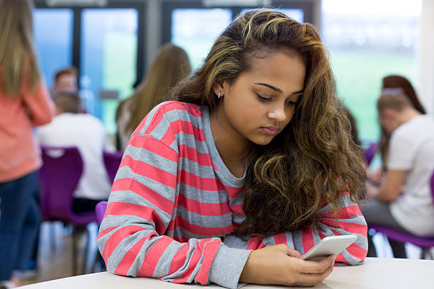 Victim of Cyber Bullying Female student sitting on her own at school. She has a smartphone in her hand and a stressed expression on her face. education student mobile phone university stock pictures, royalty-free photos & images