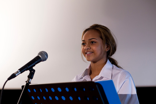 Female student making a speech. She is standing at a podium and smiling to the crowd.