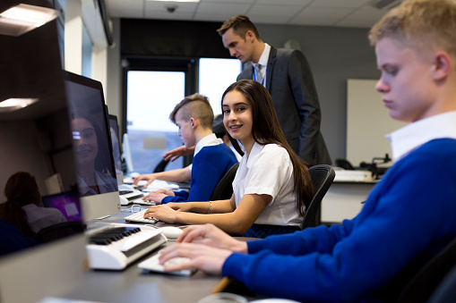 Students using computers in a school lesson. One student is smiling at the camera. There is a teacher helping another student in the background.