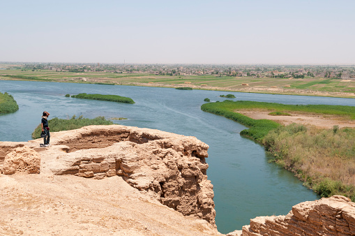 A backpacker from Japan stands overlooking the Euphrates River at Dura Europos (Tell Salhiye), an ancient ruin in eastern Syria. A town is across the river.
