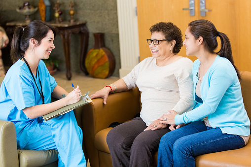 Nurse visits with Hispanic female patient and the patient's caregiver or granddaughter. The nurse is visiting the patient at home. The nurse takes notes while talking with the patient.
