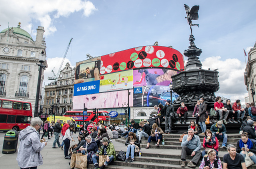 London, England - May 3, 2016: People crossing street and sit on steps of monument at Piccadilly Circus Street in London during daytime in springtime with Piccadilly Circus Shopping center behind and huge advertisement board.