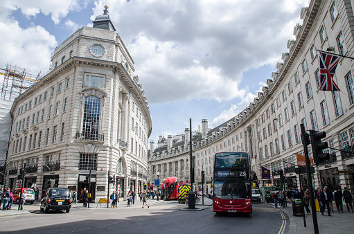 People, cars and double-decker bus passing by on Regent Street during day of springtime. There are nice building architecture behind at the curve of the street.