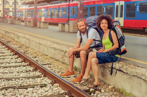 Happy young and beautiful couple of tourists with backpacks sitting at the railway station near the tracks and enjoy their journey