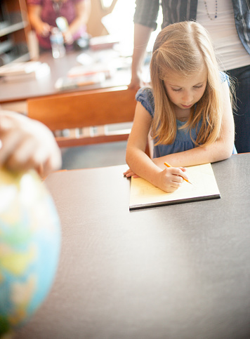 Little caucasian girl working with pencil and notepad at a desk in the library. A Globe is in the foreground.