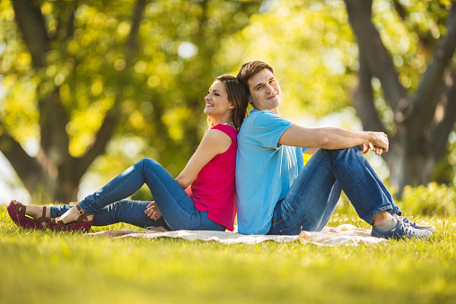 Young couple sitting back to back and enjoying a day in nature during springtime. Young man is looking at the camera.