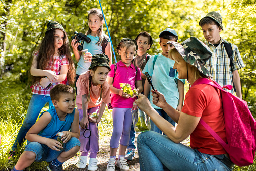 Multiracial children learning about the environment in their outdoor classroom. A 12 year old boy is sitting cross-legged on the grass taking notes. His friend, a 10 year old African-American girl, examines a leaf through a magnifying glass.