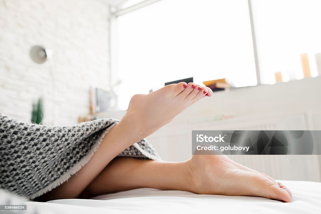 Lying on bed female legs under blanket Lying on bed female legs under blanket. Legs of young woman, that lies on her bed in bedroom under the blanket. One leg playfully lifted up. Blurred background Women Stock Photo