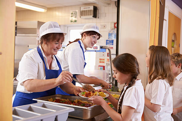 Two women serving food to a girl in a school cafeteria Two women serving food to a girl in a school cafeteria queue cafeteria worker photos stock pictures, royalty-free photos & images