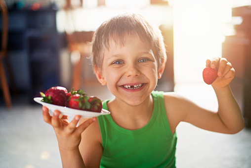 Little boy sitting on the carpet and having fun eating strawberries. Sunny day. The boy is aged 6 and is laughing at the camera holding a plate with strawberries.