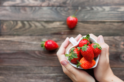 Woman holds a cup full of strawberries in hands. Mug shaped as a heart, symbol of love. Fresh juicy strawberry on rustic wooden background. Top view, place for text.