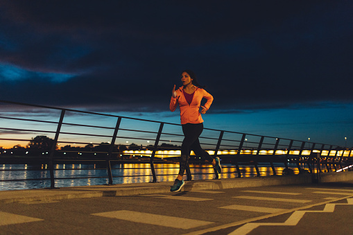 Young Woman Jogging Near River at night. She is wearing modern sporty clothing. With cityscape in background.