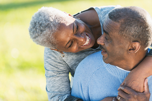 Close up of an affectionate senior African American couple outdoors.  The man is sitting, looking over his shoulder into his wife's smiling face.  She is standing behind him, bending down with her arms wrapped around him.