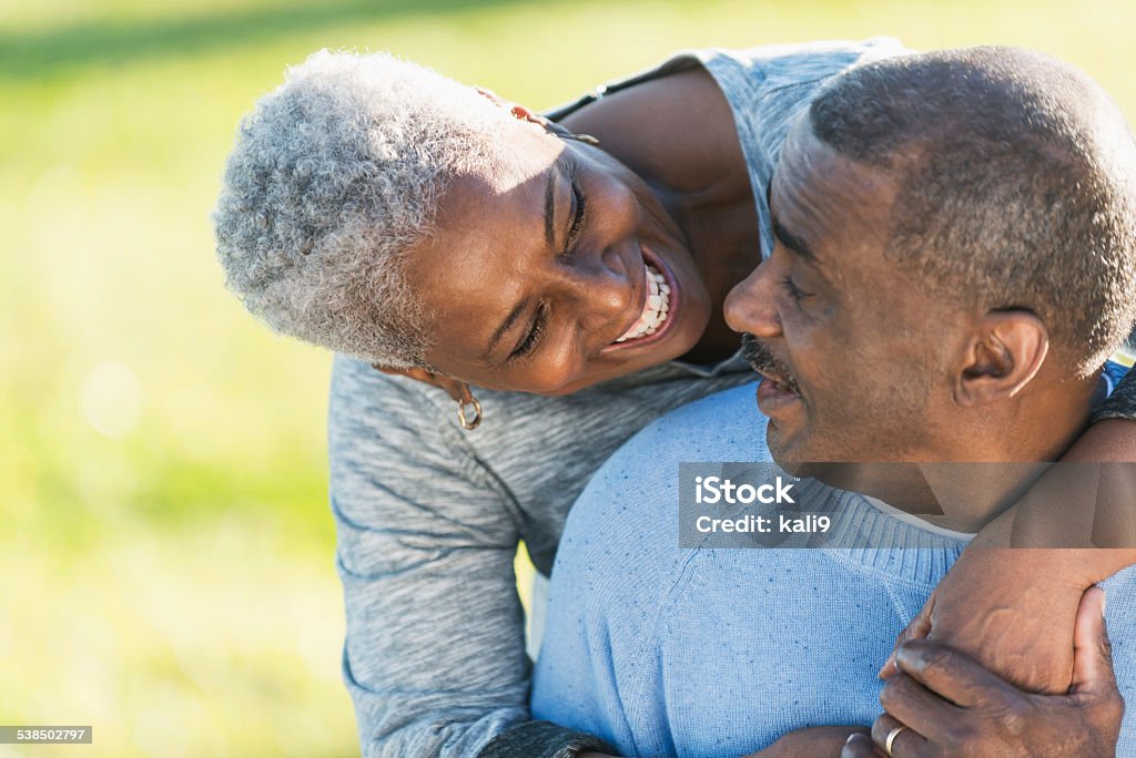 Affectionate African American couple outdoors Close up of an affectionate senior African American couple outdoors.  The man is sitting, looking over his shoulder into his wife's smiling face.  She is standing behind him, bending down with her arms wrapped around him. African-American Ethnicity Stock Photo