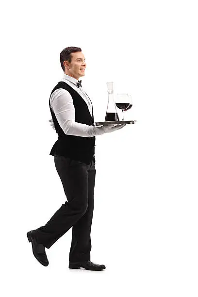 Full length profile shot of a young waiter carrying a tray with wine and two glasses isolated on white background