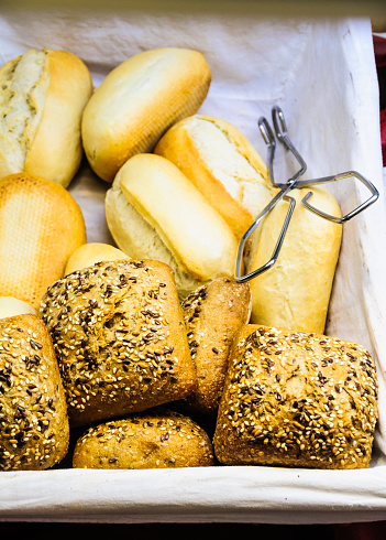 A basket of freshly baked seeded and unseeded rolls with a pair of tongs in a bakery window in the Black Forest